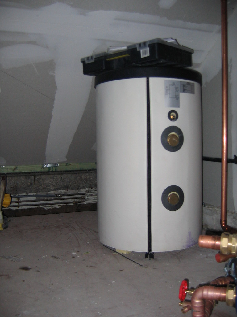 A tall factory-insulated hot-water storage cylinder in a cupboard, with pipe connections entering near the top.